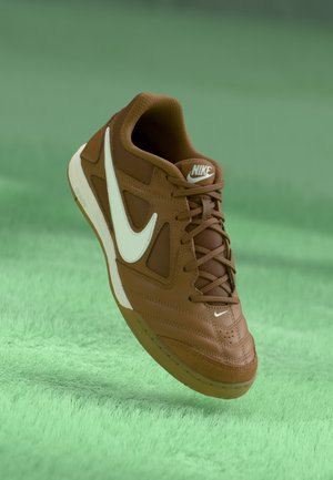 Baskets Nike marron avec logo Swoosh blanc, lacets et semelle en caoutchouc, flottant au-dessus d'une surface verte texturée.