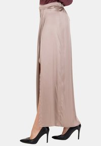 Donna che indossa una lunga gonna beige in satin con spacco laterale, abbinata a scarpe nere con tacco alto, in piedi contro uno sfondo semplice.