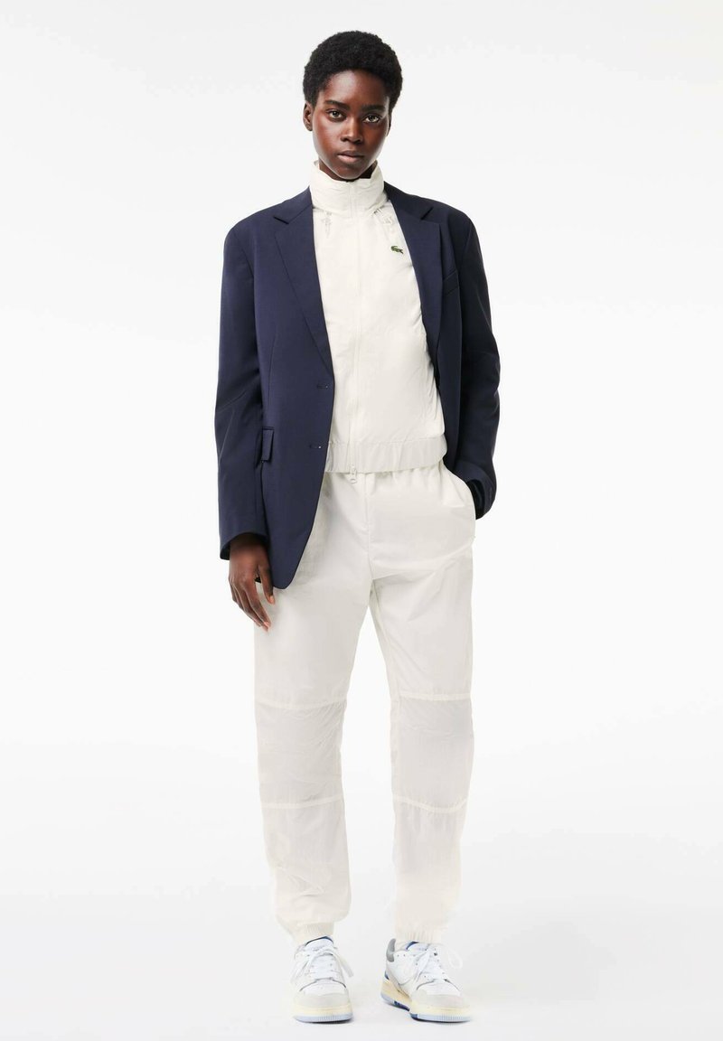 Granatowa marynarka na kremowej kurtce z wysokim kołnierzem oraz dopasowanych spodniach dresowych, z teksturowanym wykończeniem i białych trampkach z szarymi akcentami.