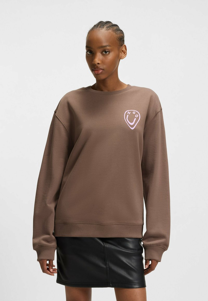 Sweat-shirt brun à manches longues et poignets côtelés, arborant un logo rose clair sur la poitrine. Associé à une jupe en cuir noire.