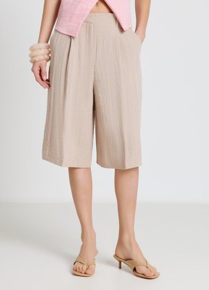 Donna che indossa pantaloncini beige con trama fino al ginocchio, top rosa chiaro senza maniche, sandali beige con tacco e braccialetti traslucidi al polso sinistro.