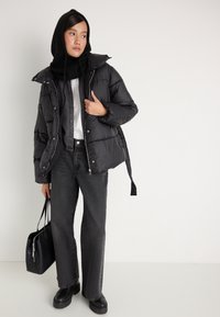 Casaco puffer preto com um acabamento brilhante, combinado com uma camisa cinza de botões e calças largas cinzas. Acompanhado de uma bolsa preta e botas.