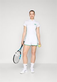 White tennis romper with green accents, short sleeves, and a zippered collar. Model holds a tennis racket and ball, wearing white socks and shoes.
