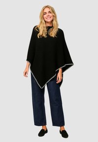 Poncho nero con texture a coste e bordi bianchi, caratterizzato da un design triangolare e morbido. Abbinato a jeans scuri a gamba larga e scarpe nere senza lacci.