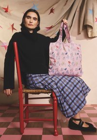 Ženska v črnem puloverju in modro karirasti krilu sedi na rdečem stolu, v rokah drži roza torbo z vzorcem in večbarvnimi ilustracijami psov.