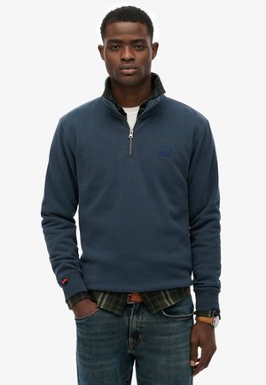Suéter de cuello alto de color azul marino con media cremallera, puños de canalé y logotipo bordado. Lleva encima una camisa de cuadros verdes, combinado con jeans azules.