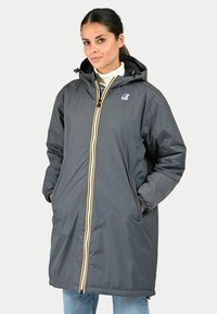 Manteau long gris avec capuche, fermeture éclair à l'avant avec des accents blancs et jaunes, comportant des poches latérales et un logo sur la poitrine. Texture lisse.