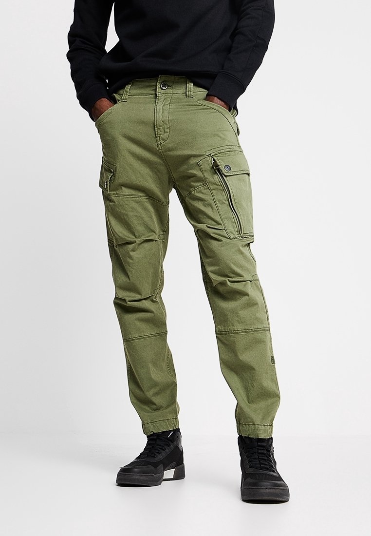 Man wearing olive green cargo pants with multiple pockets and black sneakers, hands in pockets, standing against white background.