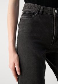 Jeans de mezclilla negra con un corte ajustado, con cintura alta, costuras en contraste y un diseño clásico de cinco bolsillos. Tejido texturizado con ligero desgaste.