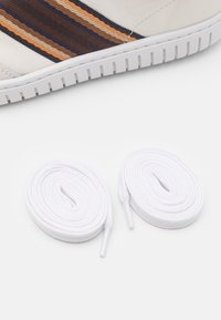 Baskets en cuir blanc avec des accents à rayures horizontales en marron, orange et marine. Deux rouleaux de lacets blancs placés à côté de la chaussure.