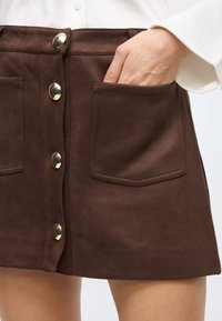 Brown suede mini skirt with a front button closure and two large pockets. Features gold-tone hardware and a smooth, soft texture.