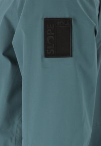 Teal fabric jacketin osa, jossa on teksturoitu musta neliöpatchi, johon on merkitty "SLOPE STYLE" kohotetulla tekstillä, esittäen mattapintaista viimeistelyä.