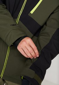 Personne ajustant la manche d'une veste O'Neill vert olive et noire avec une fermeture éclair jaune et un détail de sangle à velcro.