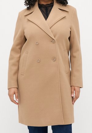 Beżowy, dwurzędowy wełniany płaszcz z szerokimi klapami, noszony na czarnej koronkowej bluzce i niebieskich dżinsach.