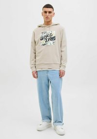 Beige hættetrøje med et blomsterdesign og sort tekst, parret med lyseblå denim og hvide sneakers. Har en hætte med snøre og ribbede manchetter.