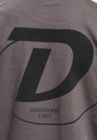 Grå skjorte med et stort sort "D" logo og teksten "WORKATION MODE F/W23" trykt på ryggen. Blødt stof med en glat overflade.