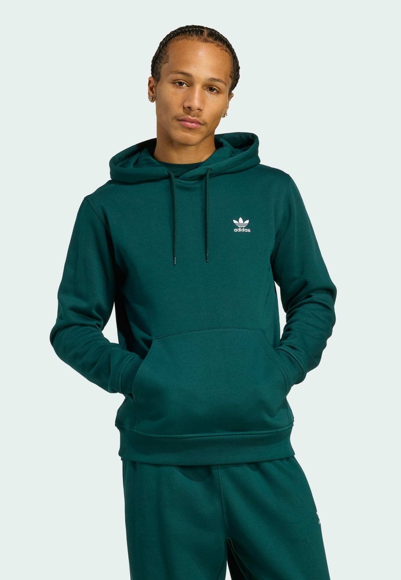 Zelená mikina s kapucňou a predným vreckom, s rebrovanými manžetami. Obsahuje biele logo adidas na hrudi. Materiál sa zdá byť mäkký a hladký.
