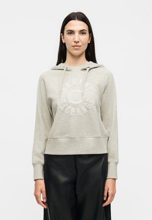 Femme aux longs cheveux foncés portant un sweat à capuche Armani Exchange gris clair et un pantalon noir, debout devant un fond blanc.
