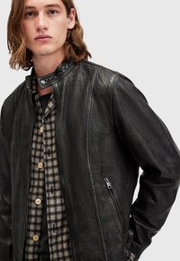Giubbotto di pelle nera con una texture liscia, colletto a scatto e dettagli di zip laterale sopra una camicia a quadri grigia e beige.