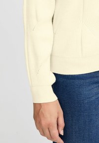 Krémový pletený svetr s texturem, s navrženými manžetami a lemem. V kombinaci s tmavě modrými džíny, které zdůrazňují kontrast mezi barvami.