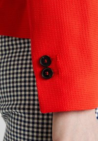 Punainen teksturoitu kangas, jossa lähikuva hihasta, jossa on kaksi mustaa nappia. Alla ruudullinen kuvio tummansinisen ja kermanvärisenä.