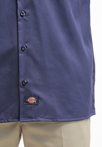 Navy blauwe button-up shirt van gladde stof, met zwarte knopen en een Dickies logo label aan de onderzoom.