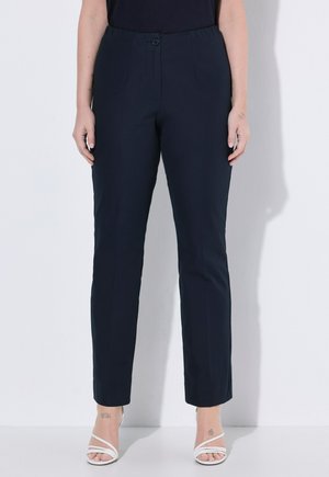 Donna che indossa pantaloni neri su misura alla caviglia con bottone e vita elastica, abbinati a sandali bianchi con cinturini.