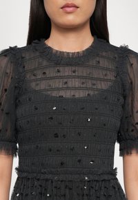 Femme portant une robe noire en tissu transparent avec un col à volants, des manches bouffantes, des rayures texturées horizontales et de petites paillettes dispersées.