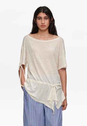 Joven con cabello largo y oscuro viste una blusa beige holgada con lazos en las mangas y pantalones anchos a rayas azules, de pie frente a un fondo liso.