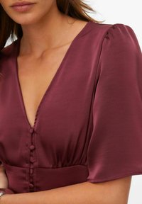 Burgundska haljina s dubokim V-izrezom, kratkim puf rukavima i gumbima s prednje strane. Glatka tkanina s lagano sjajnim izgledom.