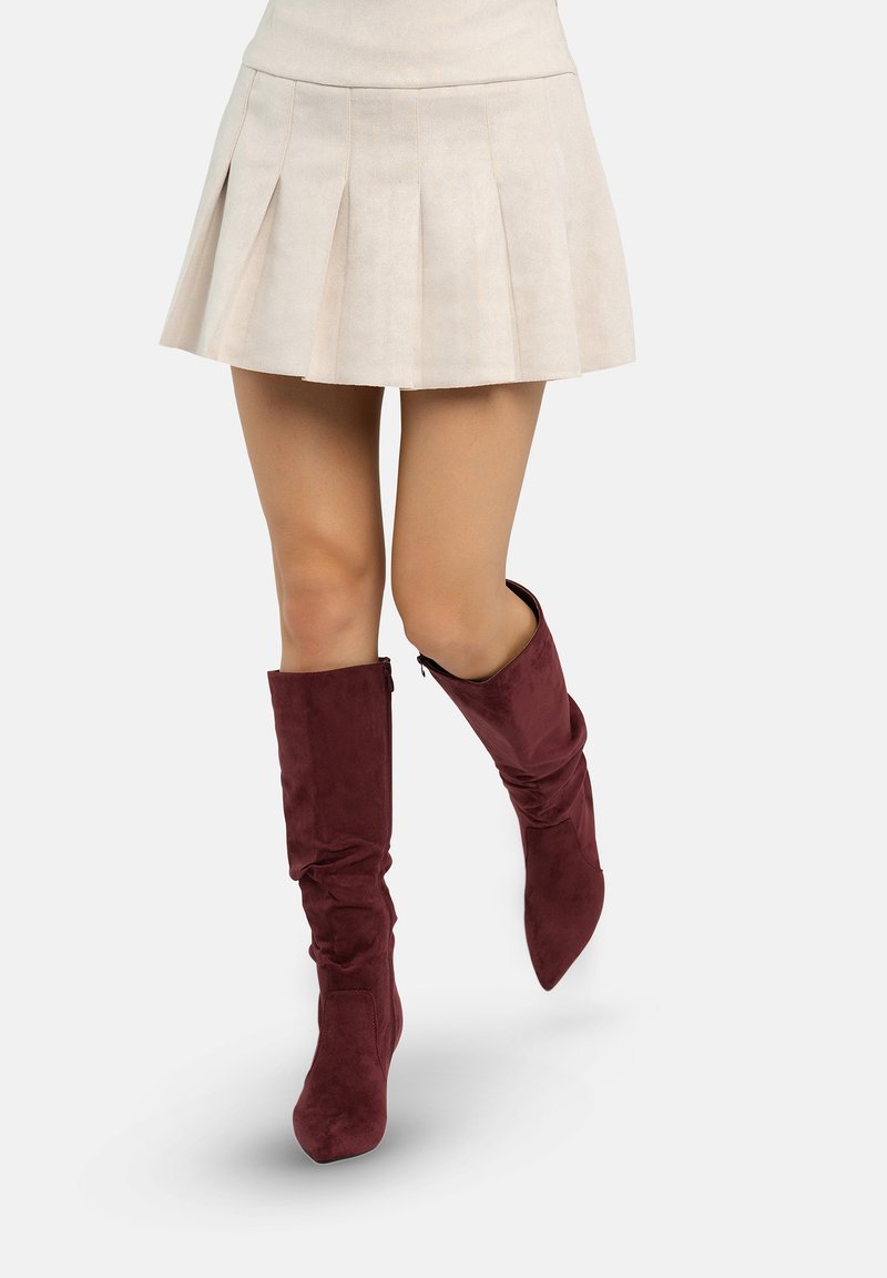 Bordeauxrode suède laarzen tot aan de knie met een slouchy ontwerp, spitse neus en een rits aan de zijkant, gecombineerd met een korte beige geplooide rok.