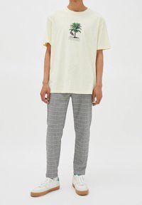 T-shirt di cotone gialla con design di palma e fenicottero. Abbinata a pantaloni a quadrati grigi e sneakers bianche con suola beige.