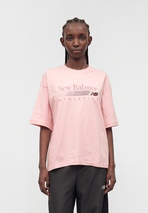 Mujer con trenzas que lleva una camiseta suelta rosa de New Balance Athletics y pantalones negros sobre un fondo claro y liso.