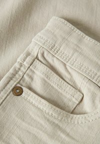 Beige bomullsstoff i nærbilde som viser en lomme med synlig søm og en bronseknapp. Teksturen er glatt med et subtilt mønster.