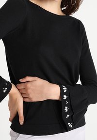 Zwarte langemouwen top met klokvormige manchetten en witte bloemenborduursels. Zacht materiaal en aansluitend ontwerp.