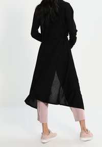 Cardigan long noir avec ceinture à la taille, fente ouverte dans le dos et design fluide. Associé à un pantalon plissé rose clair et des baskets rose doux.
