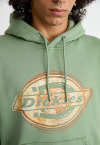 Fato de treino verde com uma grande estampa circular em tons terrosos e o texto "Dickies". O material é suave, com um ajuste padrão e cordões.
