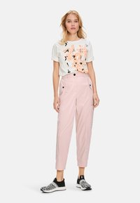 White short-sleeve t-shirt with orange graphic design, worn with light pink cargo pants featuring pockets and black accents, completed with black sneakers.