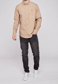 Beige overhemd van katoenen stof met lange mouwen, gecombineerd met zwarte slim-fit jeans en witte sneakers. Voorzien van een klein geborduurd logo.