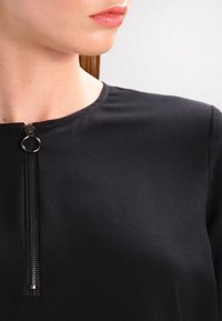 Top nero con scollo rotondo, leggero lucido e una cerniera frontale a contrasto con un tirante circolare. Design morbido e aderente.