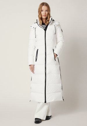 Kobieta w długim, białym, ocieplonym płaszczu z czarnym zamkiem błyskawicznym i kieszeniami, stojąca z jedną ręką w kieszeni na jednolitym tle.