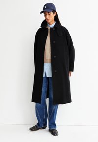 Frau, die eine marineblaue Mütze, einen übergroßen schwarzen Mantel, einen beigen Pullover, ein hellblaues Hemd, blaue Jeans und schwarze, beschlagene Slipper trägt, steht vor einer weißen Wand.