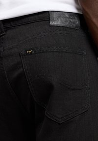 Nahaufnahme der hinteren Tasche einer schwarzen Denim-Jeans mit kleinem "Lee"-Label und einem Leder-Logo am Bund, getragen mit einem weißen Hemd.