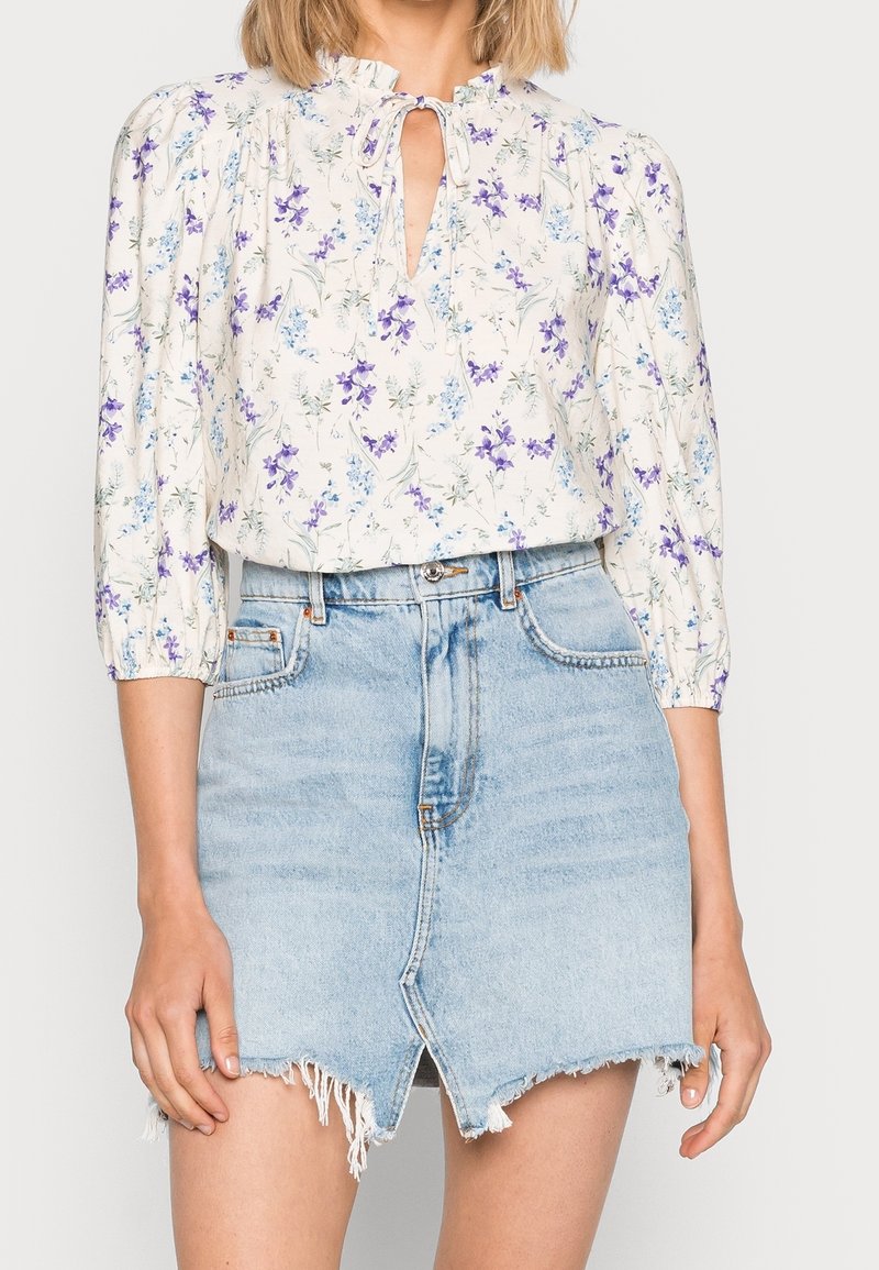 Femme portant un chemisier blanc à motifs floraux avec des manches trois-quarts et une mini-jupe en jean bleu clair avec une fente avant et un ourlet effiloché.