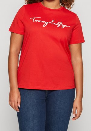 Femme portant un t-shirt rouge à manches courtes avec « Tommy Hilfiger » écrit en lettres blanches sur la poitrine et un jean bleu.