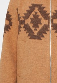 Brun ulden frakke med et geometrisk mønster i mørkere brunt, med lynlås foran, tekstureret overflade og diskrete sidelommer.