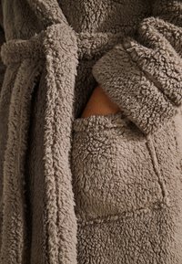 Peignoir beige duveteux avec un extérieur texturé et moelleux et une taille nouée. Comprend une poche avant et de larges poignets pour un confort accru.