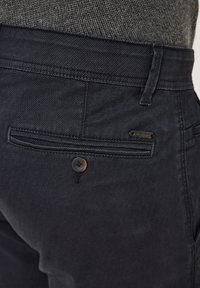 Dark navy denim pants featuring a back pocket with a button closure and a small branded tag, with a smooth texture and tailored fit.