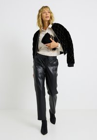 Veste bomber en velours noir avec quiltage en losange, blouse crème, pantalon en cuir noir brillant, et bottines à bouts pointus. Pochette rayée noire avec chaîne.