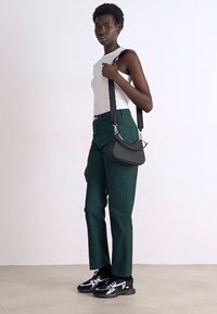 Pantalons verts ajustés, top sans manches blanc, sac bandoulière noir et baskets noires avec accents blancs. Textures lisses et design moderne.